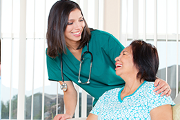 This is a picture of a nurse wearing green scrubs smiling at a female patient while having her hand rest on the patient shoulders