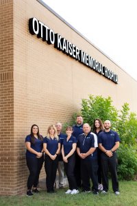 This is a picture of some of the staff for physical therapy. To the far left there is a male with a black polo shirt and tan pants on next to him is a female with long brown hair with blue scrubs on next to her is another female with brown hair behind her is a male with a blue polo on next to him is a older lady with green scrubs and a blue long sleeve on next to her is a lady with green scrubs and to the far right there is a male with blue scrubs on